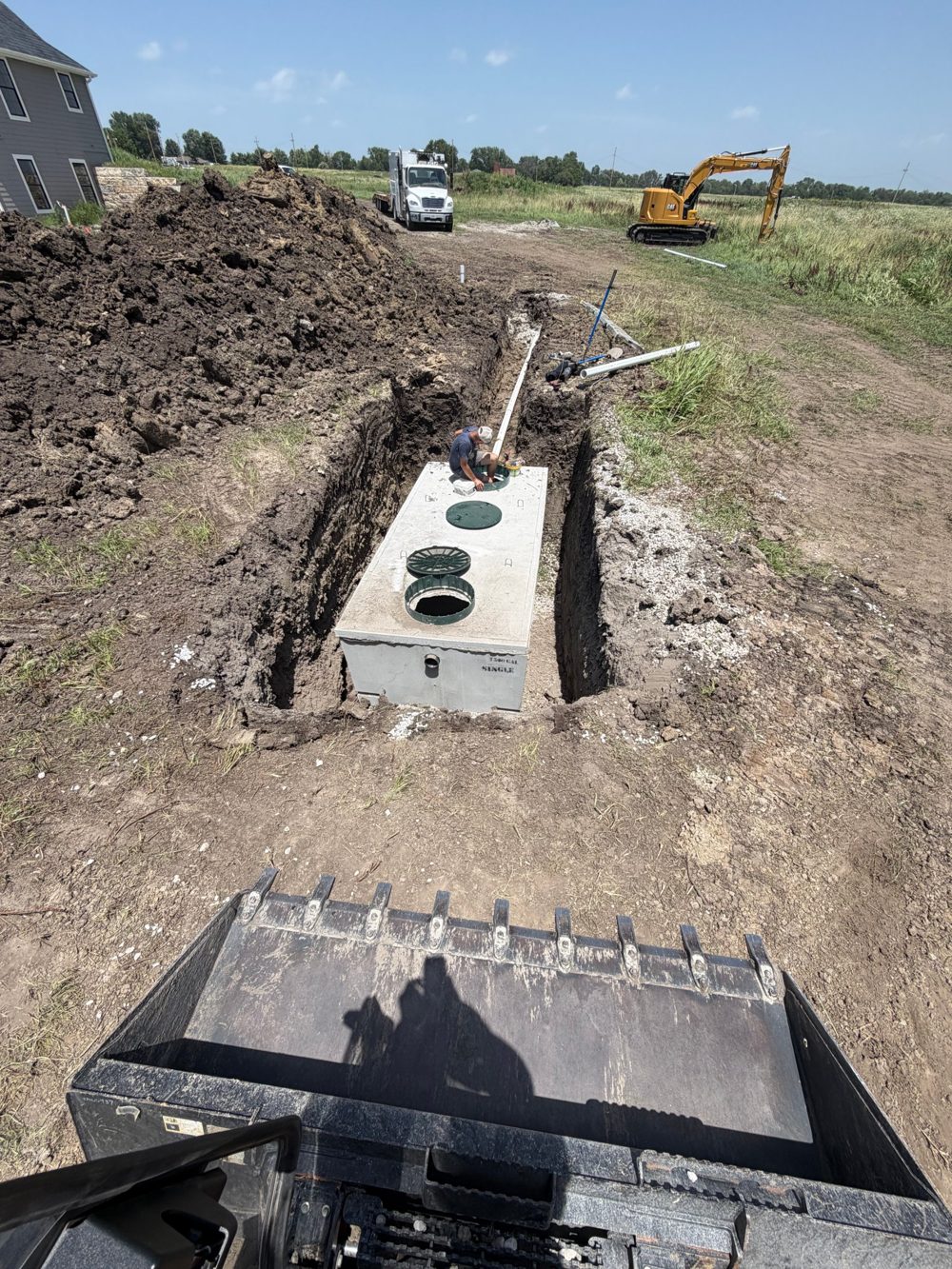A new concrete septic tank set in an excavated trench beside a new home, with an excavator and work truck on site