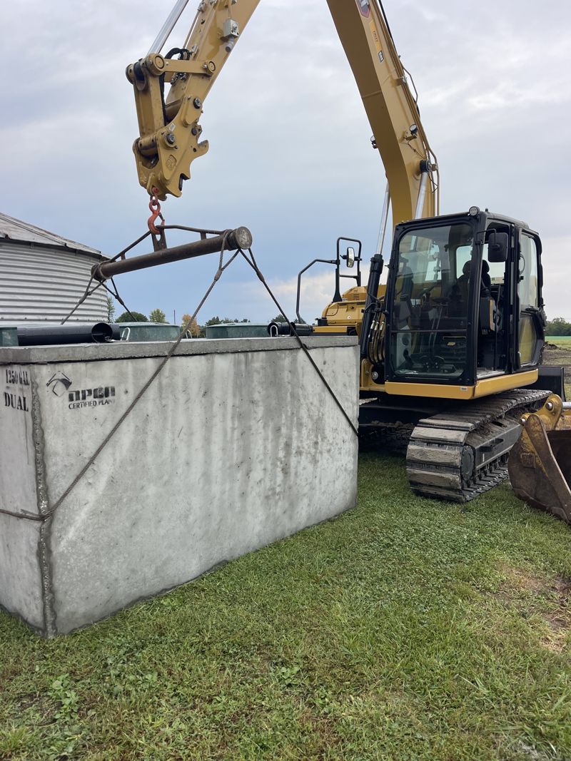 Excavator lifting a 1,250 gallon concrete septic tank with a spreader bar