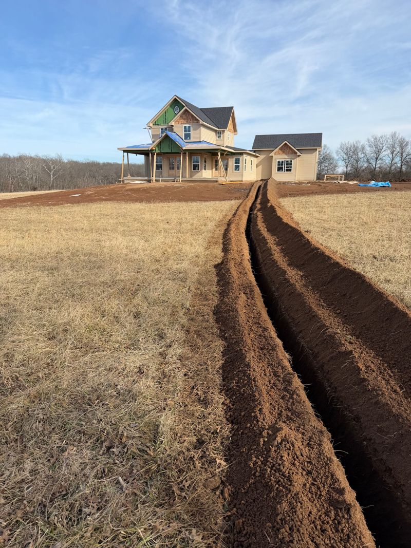 Long straight trench in red soil leading across a field to a new house under construction
