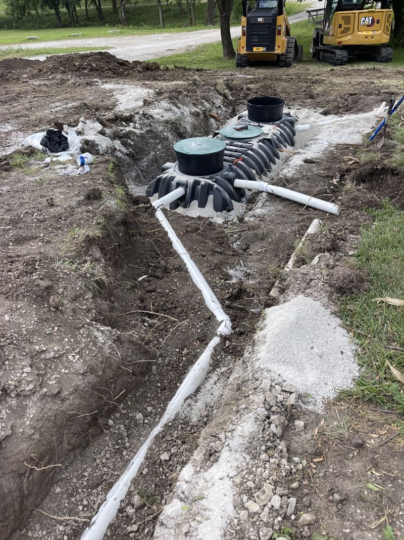 Black plastic septic tank with green lids and white PVC lines in an open excavation