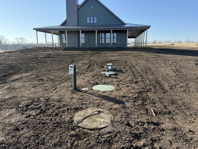 Septic lids, riser and control panel post in freshly graded dirt in front of a green metal house with a wraparound porch